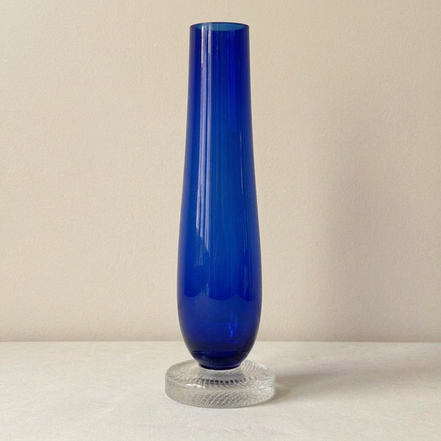 Jarrón azul recto con pie de cristal sobre piedra clara y pared beige